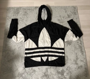 Vindjacka från Adidas - Svart och vit vindjacka från Adidas med stor Trefoil-logga över hela jackan. Jackan har huva, dragkedja framtill och meshfoder på insidan. Perfekt för dig som vill sticka ut med en sportig och streetig stil.