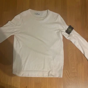 Vit långärmad tröja från Stone Island - Vit långärmad tröja från Stone Island med klassisk rund hals och ikonisk svart patch med broderad logga på vänster ärm. Tröjan är tillverkad i mjuk bomull och har en clean, stilren look som passar perfekt till jeans eller cargo. Snygg och enkel att matcha.