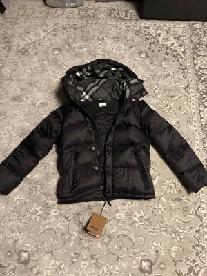 Svart pufferjacka från Burberry - Snygg svart pufferjacka från Burberry med klassiskt rutigt foder i grått och vitt. Jackan har huva, dragkedja och knappar framtill samt två fickor med dragkedja. Stor Burberry-patch på ärmen ger en exklusiv vibe. Perfekt för kalla dagar.