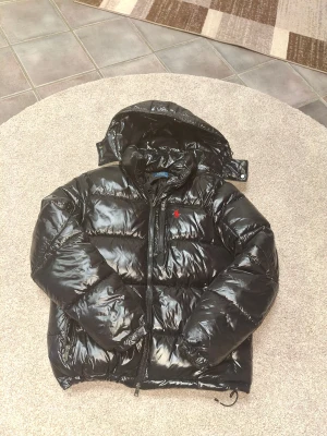 Svart pufferjacka Polo Ralph Lauren - Svart glansig pufferjacka från Polo Ralph Lauren med röd broderad logga på bröstet. Jackan har huva, dragkedja framtill och två sidofickor med dragkedja. Perfekt för kalla dagar och har en riktigt modern vibe.