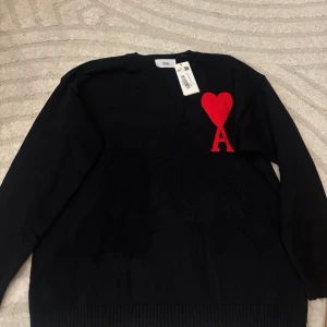 Svart stickad tröja från Ami Paris - Svart stickad tröja från Ami Paris med rött hjärta och bokstaven A broderat på bröstet. Tröjan har rund hals, ribbade muddar och lång ärm. Perfekt för dig som gillar stilrena plagg med en twist. Snygg detalj framtill som gör den unik. Inte äkta kommer med taggen osv.