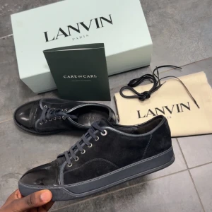 Svarta sneakers från Lanvin i mocka - Snygga svarta sneakers från Lanvin med ovandel i mocka och detaljer i blankt skinn på tån. Skorna har svarta snören, platt sula och diskret branding. Perfekta för dig som gillar stilrena och lyxiga sneakers med en clean look.