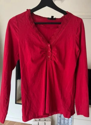 Röd blus med spets från KappAhl - Snygg röd blus från KappAhl i storlek S med lång ärm och v-ringad hals. Blusen har dekorativ spets runt halsringningen och knappar framtill. Materialet är mjukt och lätt, perfekt för dig som gillar detaljer och färg.