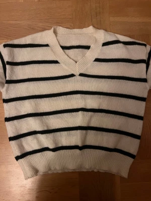 Vit och svart randig stickad tröja - Snygg stickad tröja med svarta ränder på vit botten och v-ringad hals. Modellen har ribbade muddar och finns både med lång ärm och som ärmlös variant. Perfekt för dig som gillar klassisk stil med en twist.