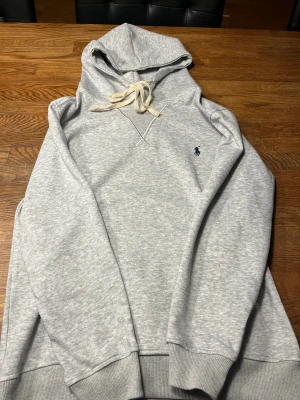 Grå hoodie från Polo Ralph Lauren - Säljer på grund av fel storlek. Snygg ljusgrå hoodie från Polo Ralph Lauren med klassisk blå logga på bröstet. Tröjan har huva med snörning, ribbade muddar och är tillverkad i mjuk bomullsmix. Perfekt för chill dagar och enkel att matcha med jeans eller joggers.