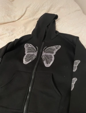 Svart hoodie med fjärilstryck från SHEIN - Svart hoodie från SHEIN i storlek M med stora vita fjärilar tryckta på bröstet och flera mindre fjärilar längs ena ärmen. Tröjan har huva, dragkedja framtill och praktiska fickor. Materialet är mjukt och känns som polyester, perfekt för chill dagar.