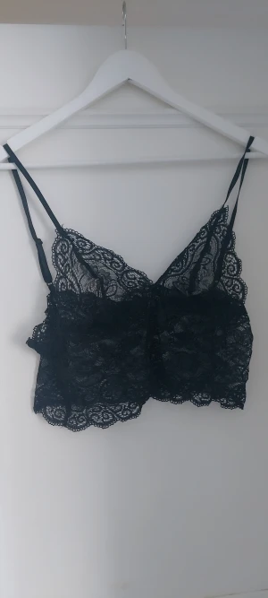 Svart spetsig linnetopp med tunna band - Säljer en svart linnetopp i spets med tunna, justerbara axelband. Toppen har v-ringad form och är croppad med fina spetsdetaljer längs kanterna. Perfekt för att ge outfiten en extra touch av stil.