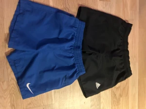 Nike och Adidas Badshorts - Två par sportiga shorts, ett par blå från Nike med vit logga och ett par svarta från Adidas med vit logga. Båda har elastisk midja och är tillverkade i lätt syntetmaterial, perfekta vad en varm dag! 1 Par för 100, 2 för 175:-