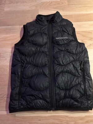 Svart dunväst från Peak Performance - Svart dunväst från Peak Performance med quiltad design och hög krage. Västen har två dragkedjeförsedda fickor på sidorna och logga på bröstet. Perfekt för lager-på-lager och enkel att matcha med olika outfits. Nypris 1599:-