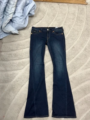 True Religion Joey Low Rise Flare Jeans - Snygga mörkblå jeans från True Religion, modell Joey Low Rise Flare. De har låg midja, klassiska fem fickor och utställda ben med coola kontrastsömmar. Bakfickorna har den ikoniska hästskosömmen. Perfekta för dig som gillar retrovibbar och statement-jeans.