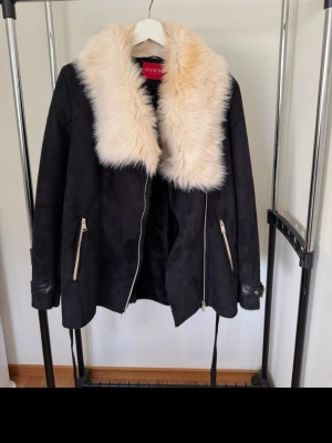 Svart pälsjacka från Guess - Snygg svart pälsjacka från Guess med stor fluffig beige krage i fuskpäls. Jackan har dragkedja framtill, två snedställda fickor med dragkedjor och detaljer i skinnimitation vid ärmsluten. Perfekt för dig som vill ha en statementjacka med lyxig känsla.