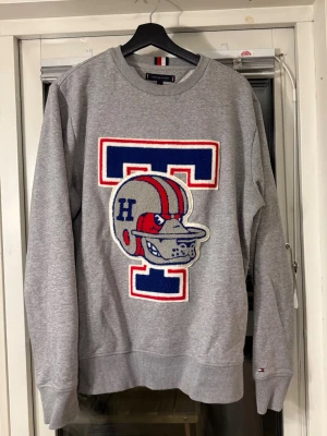Grå college-tröja Tommy Hilfiger - Grå sweatshirt med stort blått och rött T-tryck och en bulldog med hjälm på bröstet. Klassisk rund hals och ribbade muddar. 