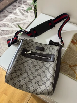 Beige axelväska med monogram - Snygg axelväska  i beige och brunt. Väskan har bruna skinnkanter, en dragkedjeficka framtill och en bred axelrem i rött och marinblått. Perfekt för dig som vill ha en stilren och trendig väska.