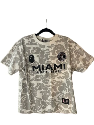 BAPE x Inter Miami camo t-shirt - Snygg t-shirt från BAPE i samarbete med Inter Miami. T-shirten har grått och vitt camomönster, tryck med MIAMI och BAPE-logga fram, samt stort BAPE 93-tryck bak. Korta ärmar och rund hals. Perfekt för dig som gillar streetwear och unika samarbeten. Liten i storleken. 