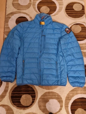 Blå dunjacka från Parajumpers - Snygg blå dunjacka från Parajumpers med hög krage och dragkedja framtill. Jackan har två sidofickor med dragkedja och en patch på ena ärmen. Perfekt för kyliga dagar och enkel att matcha med olika outfits. Är lite sönder kolla bild 4. Är öppet vid armhålorna men det är desingen.