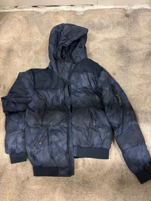 Mörkblå pufferjacka med huva - Snygg mörkblå pufferjacka från Etnies med huva och dragkedja framtill. Jackan har diskret mönster, ribbade muddar och fickor med knappstängning. Perfekt för kalla dagar när du vill hålla stilen.