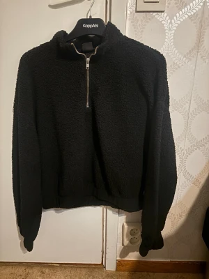 Svart teddytröja från Gina Tricot - Mysig svart teddytröja från Gina Tricot med halv dragkedja framtill och hög krage. Tröjan har långa ärmar och resår i midjan, perfekt för kalla dagar. Materialet är fluffigt och ger en cool, avslappnad look.