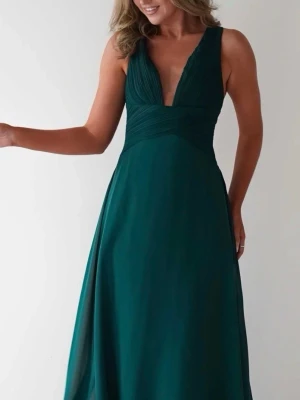 Mörkgrön långklänning med v-ringning - Elegant mörkgrön långklänning med djup v-ringning och korsade axelband i ryggen. Klänningen har en figurnära överdel och ett svepande, lätt utställt kjolparti. Tillverkad i ett mjukt, flytande material som ger en lyxig känsla.
