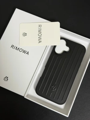 Svart iPhone 16 Rimowa mobilskal - Stilrent svart mobilskal från Rimowa med räfflad design och diskret logotyp. Skalets form är anpassad för iPhone och har ett robust yttre i hårdplast som skyddar mobilen. Perfekt för dig som vill ha ett exklusivt och modernt tillbehör till din telefon.