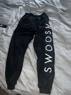 Nike SWOOSH nylon - Säljer ett par svarta mjukisbyxor från Nike med stort vitt SWOOSH-tryck längs ena benet och Nike-logga på höften. Byxorna har resår i midjan och vid bensluten samt snörning i midjan. Perfekta för chill eller streetwear.