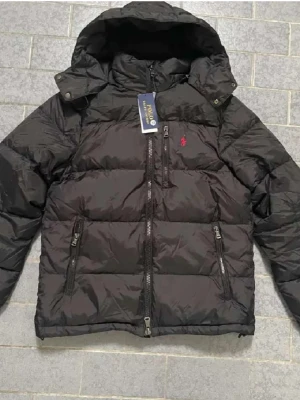 Svart pufferjacka från Polo Ralph Lauren - Säljer en svart pufferjacka från Polo Ralph Lauren med huva och flera dragkedjeförsedda fickor. Jackan har det klassiska röda Polo-logot på bröstet och är perfekt för kalla dagar. Tillverkad i syntetmaterial och dunfylld för extra värme. Kontakta mig vid intresse! 