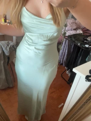 Ljusgrön långklänning i satin - Säljer en elegant ljusgrön långklänning i satin med smala axelband och figurnära passform. Klänningen har en mjukt draperad överdel och slits i nederkanten för extra rörelsefrihet, dessutom har den blommor som hänger längsmed ryggen. Perfekt för dig som vill ha en stilren och trendig look! Använd endast 1 gång på min konfirmation, har sedan dess tvättats och hängt i min garderob. ORGINAL PRIS 800.