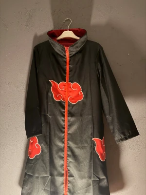 Akatsuki cosplaykappa Naruto - Svart cosplaykappa med röda moln och röd dragkedja, inspirerad av Akatsuki från Naruto. Perfekt för anime- och mangaentusiaster som vill klä ut sig till en medlem i Akatsuki. Tillverkad i syntetmaterial med ståkrage och hel dragkedja framtill. - Allting samma pris med mask också - Endast Anime älskare (Storlek: M/L