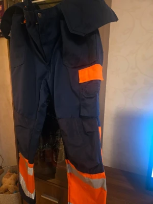 Arbetsbyxor - Säljer ett par rejäla och slitstarka arbetsbyxor från kvalitetsmärket Fristads. Byxorna är i marinblått med varseldetaljer (orange och reflex) på benen för hög synlighet och säkerhet.  Storlek: D120 passar dig som har ett midjemått på ca 120 cm. Modellen har Fristads 'D-passform', vilket innebär att benen är något kortare (innerben ca 78 cm) för att slippa att byxorna korvar sig vid fötterna."  D-storlek" (till skillnad från en C-storlek) är designad för personer som behöver mer vidd kring midja