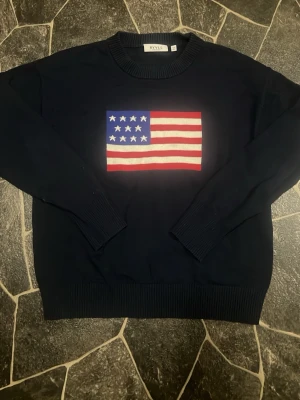 Mörkblå stickad tröja med flagga - Mörkblå stickad tröja från Ralph Lauren med en stor amerikansk flagga på bröstet. Tröjan har rund halsringning, långa ärmar och ribbstickade muddar vid ärmslut och nederkant. Perfekt för dig som gillar klassisk och stilren design.