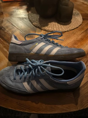 Adidas Spezial blå sneakers - Snygga Adidas Spezial sneakers i ljusblå mocka med vita ränder och detaljer. Klassisk retrostil med platt sula och snörning. Perfekta för dig som gillar streetwear och vill sticka ut med en fräsch färg på fötterna.