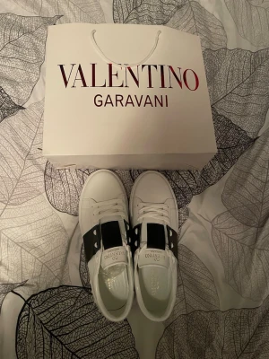 Valentino Garavani vita sneakers - Snygga vita sneakers från Valentino Garavani med en bred svart rem över ovandelen och coola nitar på hälen. Skorna har klassisk snörning och är tillverkade i skinn för en lyxig känsla. Perfekta för dig som gillar stilrena och exklusiva sneakers. Har tyvärr ingen låda eftersom jag tappat bort den men påsen är kvar. Storlek 44.