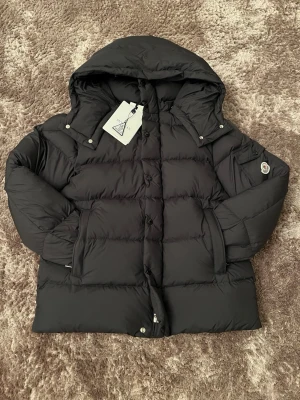 Svart dunjacka från Moncler - Snygg svart dunjacka från Moncler med huva och klassisk pufferdesign. Jackan har dragkedja och knappar framtill, två stora fickor och Moncler-logga på ärmen. Perfekt för kalla dagar med sin varma och fluffiga känsla.