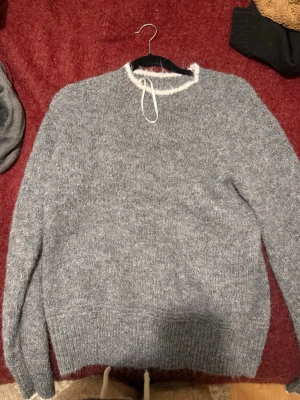 Grå fluffig stickad tröja - Mysig grå stickad tröja med fluffig känsla och vita detaljer vid halsringning och ärmslut. Tröjan har rund hals och långa ärmar, perfekt för kalla dagar. Materialet ser ut att vara en mjuk ull- eller mohairblandning. Aldrig använd