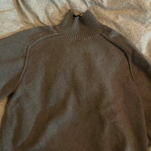 Grå/beige stickad polotröja - Mysig grå/beige stickad polotröja med hög ribbad krage och långa ärmar. Tröjan har en enkel och stilren design med ribbade muddar vid ärmslut och nederkant. Perfekt för kalla dagar och enkel att matcha med olika outfits. Färgen kommer inte riktigt fram i bilden men den är grå/beige