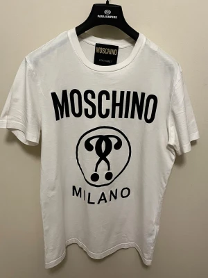 Vit Moschino Milano t-shirt - Snygg vit t-shirt från Moschino med stort svart tryck framtill där det står 'Moschino Milano' och en ikonisk logga. Klassisk rund halsringning och korta ärmar. Tillverkad i mjuk bomull för en skön känsla och clean look.