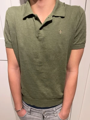 Olivgrön stickad pikétröja - Snygg merinoull olivgrön stickad pikétröja från Morris med korta ärmar och klassisk krage. Tröjan har en liten broderad logga på bröstet och ribbade muddar vid ärmslut och nederkant. Perfekt för en avslappnad men stilren look.