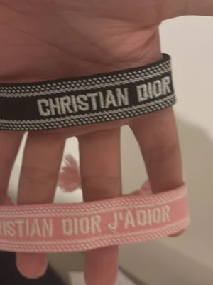 Christian Dior tygarmband 2-pack - Två snygga tygarmband från Christian Dior, ett i svart med vit text och ett i ljusrosa med vit text där det står 'CHRISTIAN DIOR J'ADIOR'. Perfekta accessoarer för att lyfta din outfit med en lyxig touch.