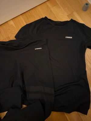 Svart träningsset från Stronger - Säljer ett svart träningsset från Stronger med matchande t-shirt och tights. Tightsen har diskreta meshdetaljer på benen och båda plaggen har Stronger-loggan tryckt i vitt. Perfekt för gymmet eller löprundan.