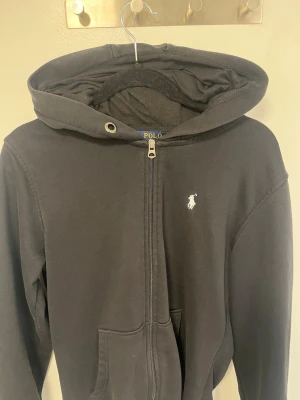 Svart hoodie från Polo Ralph Lauren - Svart hoodie med dragkedja från Polo Ralph Lauren. Klassisk design med vit broderad logga på bröstet, huva med snörning och känguruficka framtill. Tillverkad i mjukt bomullsmaterial, perfekt för en avslappnad och stilren look.