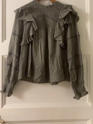 Grå volangblus med lång ärm - Stilren grön blus med volangdetaljer framtill och på axlarna. Blusen har långärmade ärmar med smock vid ärmslut och dekorativa spetsband längs ärmarna. Bara att fråga ifall du undrar något eller vill ha fler bilder! Pris kan diskuteras💞