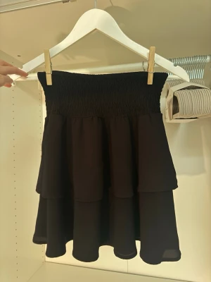 Svart volangkjol med smock - Supersnygg svart kortkjol med bred smock i midjan och dubbla volanger. Kjolen har en luftig och flowy känsla, perfekt för att styla med crop tops eller stickade tröjor. Materialet känns lätt och bekvämt.👀