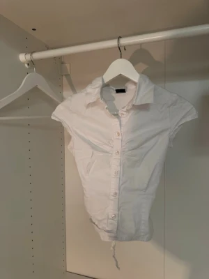 Vit croppad skjorta med knytband - Säljer denna populära och underbart vackra vita croppade skjortan med korta ärmar och knappar framtill. Skjortan har en klassisk krage och knytband i midjan för en snygg passform i midjan. Perfekt till jeans eller kjol för en trendig look. Har tyvärr blivit för liten för mig😓🤍