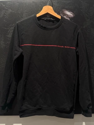 Svart sweatshirt från Prada Milano - Svart sweatshirt från Prada Milano med en tunn röd linje och diskret logga på bröstet. Tröjan har rund halsringning, långa ärmar och ribbade muddar. Perfekt för en stilren och trendig look.
