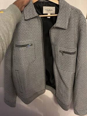 Mönstrad jacka från River Island - Säljer en svartvit jacka från River Island med cirkelmönster och dragkedjor på bröstfickorna. Jackan har krage, ribbade muddar och en klassisk passform. Perfekt för dig som vill sticka ut med en unik look.