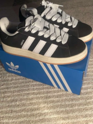 Adidas Samba svartvita sneakers - Klassiska Adidas Samba sneakers i svart mocka med vita detaljer och de ikoniska tre ränderna på sidan. Skorna har vit sula, rund tå och breda vita skosnören. Perfekta för dig som gillar retrostil och streetwear.
