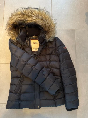 Svart dunjacka Tommy Hilfiger - Snygg svart dunjacka från Tommy Hilfiger med quiltad design och avtagbar huva med fuskpäls. Jackan har långa ärmar, dragkedja och klassisk logga på ärmen. Perfekt för kalla dagar och riktigt varm.