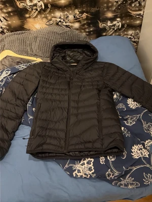 Svart dunjacka från J.Lindeberg - Snygg svart dunjacka från J.Lindeberg med huva och dragkedja framtill. Jackan har en quiltad design och är tillverkad i lätt, syntetmaterial som håller värmen. Perfekt för kalla dagar och varma dagar och enkel att matcha med olika outfits. 