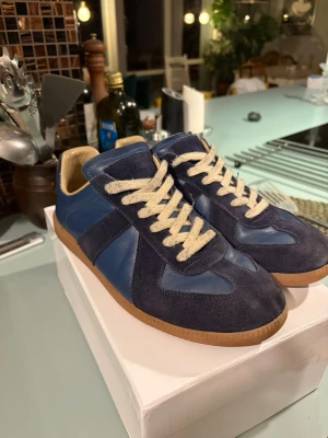 Blå maison margiela gats - Snygga sneakers i mörkblå mocka och blått skinn med beige snörning och gummisula. Skorna har en klassisk låg profil och rund tå, perfekt för en stilren och avslappnad look. Passar dig som gillar retrovibbar och tidlös design. Maison margiela gats 22, storlek 43. 8/10 skick
