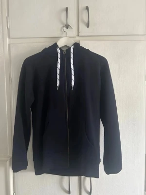 Mörkblå zip-hoodie med vita snören - Står large men skulle säga att den är mer åt medium hållet👍En stilren mörkblå hoodie med dragkedja och vita snören i huvan. Hoodien har känguruficka framtill och ribbade muddar vid ärmslut och nederkant. Perfekt för en avslappnad look.