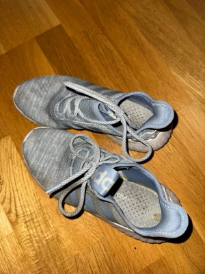 Ljusblå Adidas sneakers med snörning - Säljer ett par ljusblå Adidas sneakers med klassiska tre ränder på sidan och snörning. Ovandelen är i textil och syntetmaterial, och sulan är vit och räfflad för bra grepp. Perfekta för dig som gillar sportig och avslappnad stil.